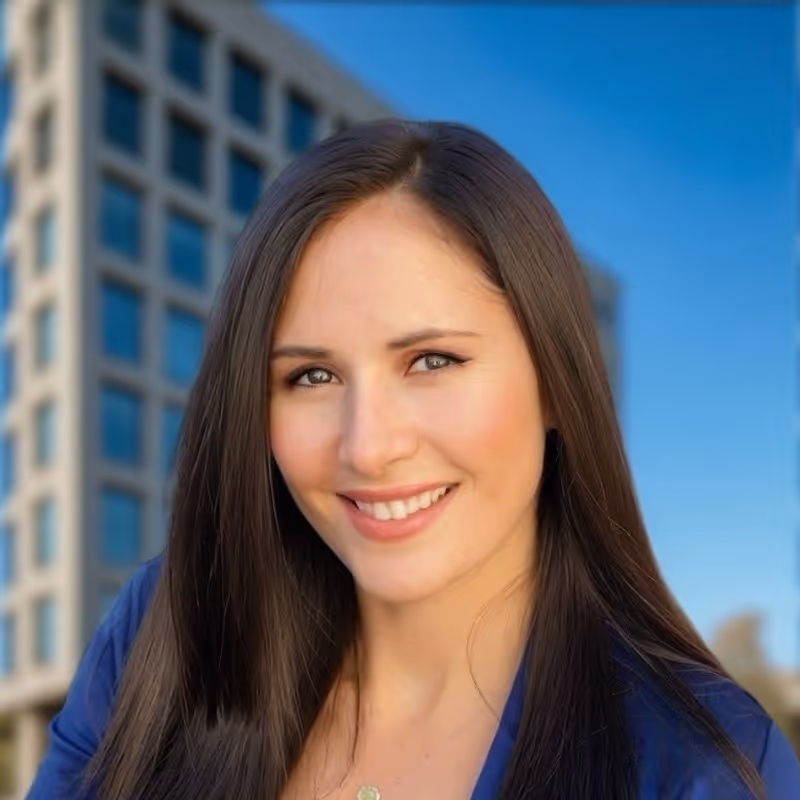 Smiling woman with long brown hair wearing a blue top against a background of a modern building and clear sky.