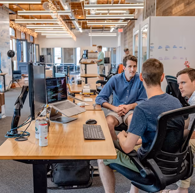 Three men having a discussion around a desk with computer monitors and laptops in a modern open-plan office.