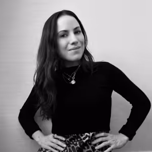 Black-and-white portrait of a woman with long dark hair wearing a black turtleneck and a patterned skirt, smiling with hands on hips.