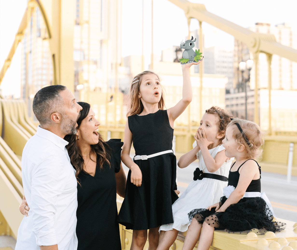 Family of five on a bridge, with a young girl holding a small cartoon rhinoceros above her head as others look surprised.