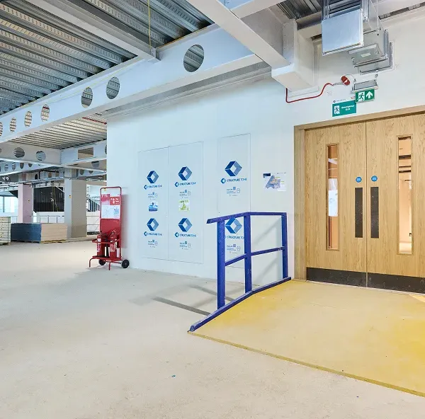 Corridor under refurbishment with protective flooring, a blue barrier and timber double doors.
