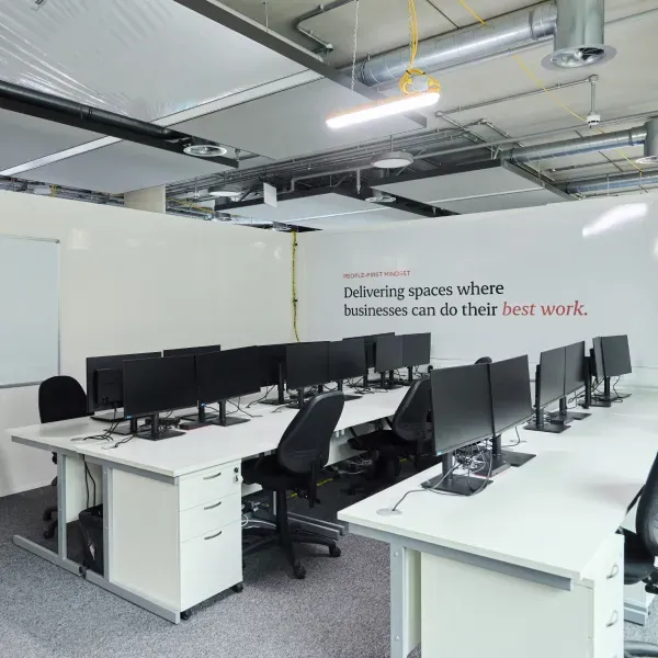Open-plan office with desks, monitors and exposed ceiling services.