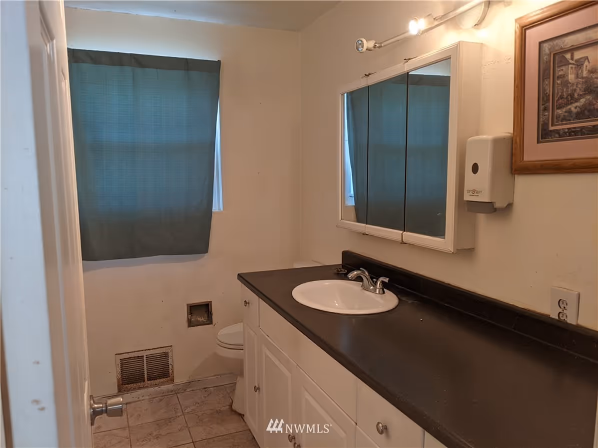 Small outdated bathroom with dark countertop, single sink, basic mirror cabinet, and a curtained window.