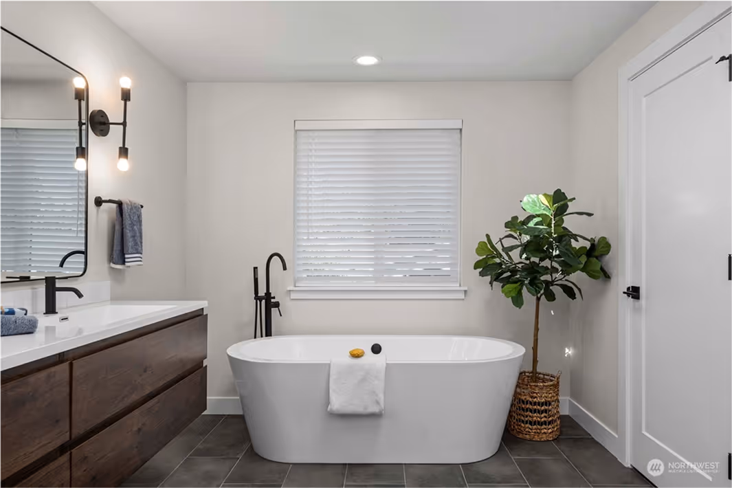 Modern bathroom with a freestanding white soaking tub, matte black floor-mounted faucet, and a large window with white blinds. A potted plant sits beside the tub on dark tile flooring.