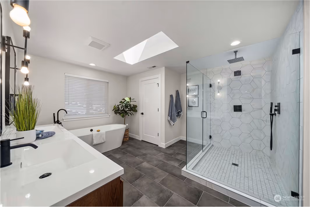 Spacious bathroom featuring a freestanding tub, large walk-in glass shower with geometric tile, dark tile floors, and a floating wood vanity with double sinks.