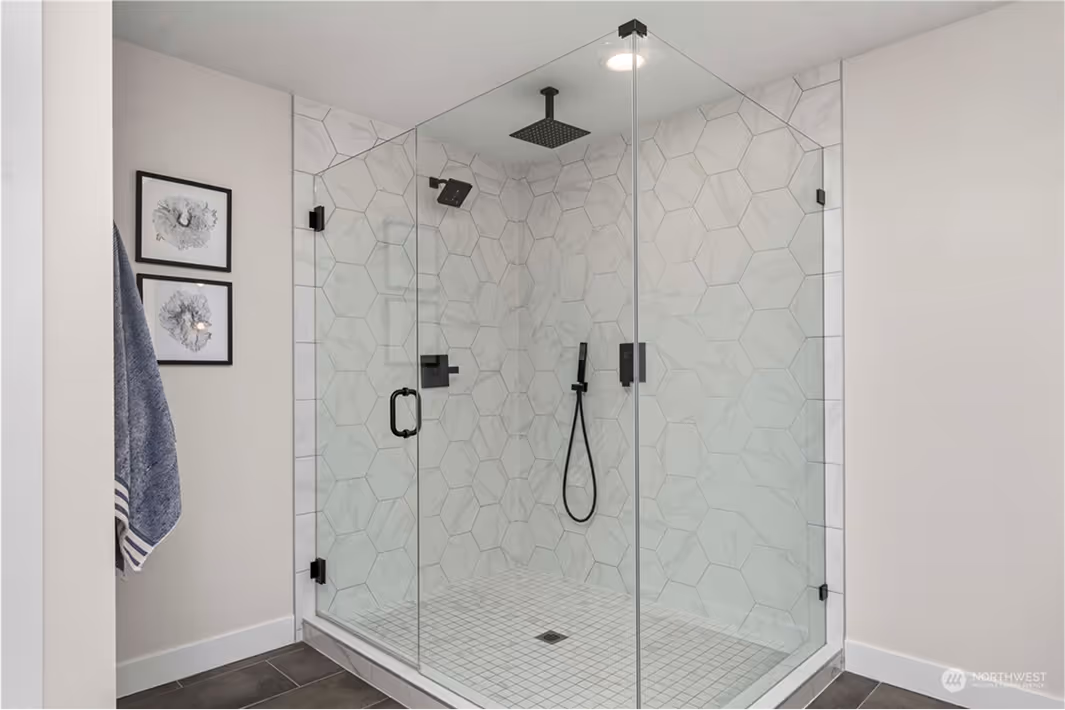 Frameless glass walk-in shower with large geometric hex tiles in a marble pattern, matte black shower fixtures, and modern lighting.