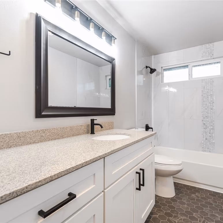 Full bathroom view with granite countertop, white shaker vanity, black fixtures, and tiled shower with decorative accent.