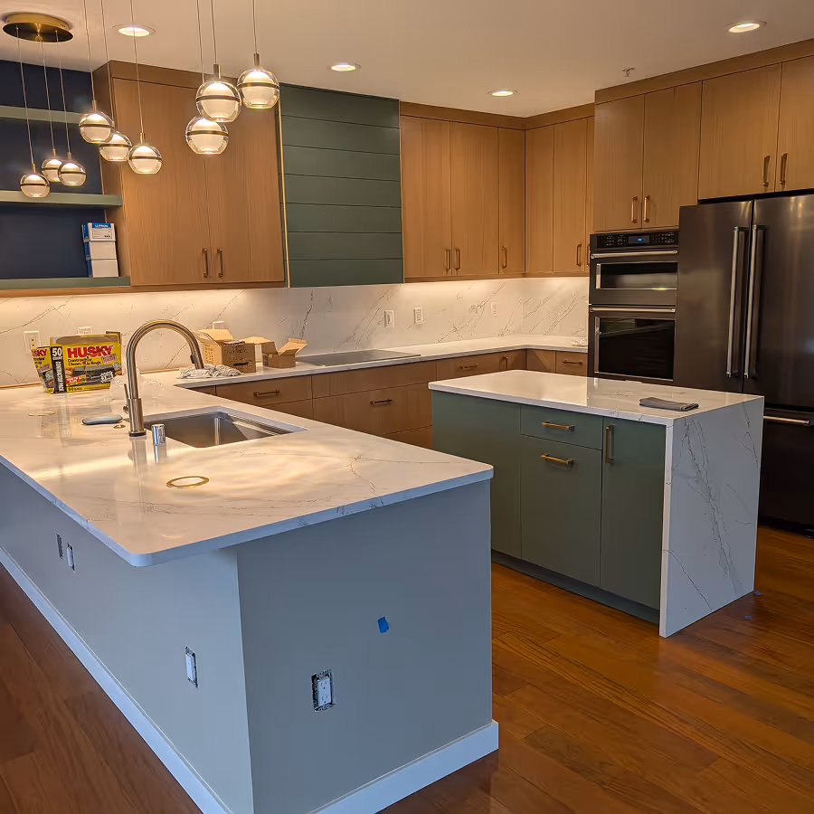 Spacious modern kitchen with warm wood cabinets, two islands with white counters, green accents, and hanging globe pendant lights.