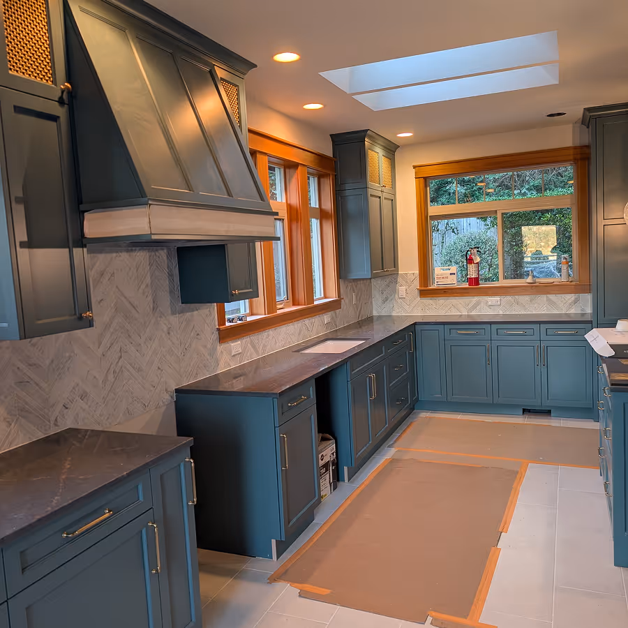 Kitchen renovation featuring deep teal cabinets, a matching large range hood, white herringbone tile backsplash, and wood window trim.