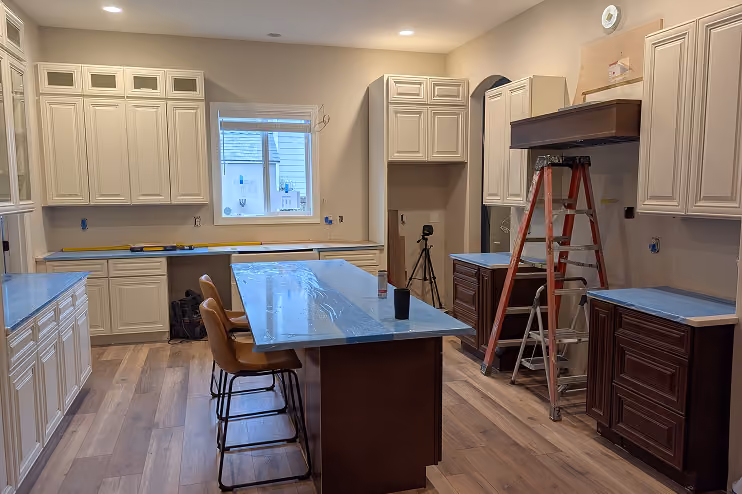 Kitchen under renovation with white and dark wood cabinets, a central island with plastic-covered counters, two brown stools, a red ladder, and some tools on the floor and counters.