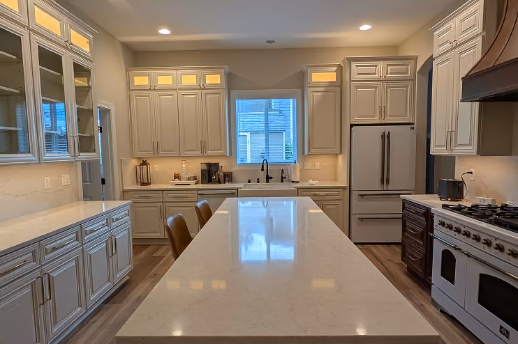 Modern kitchen with a long central island featuring a glossy white countertop and two brown bar stools, cream-colored cabinets with glass panels on the left, a window above the sink at the back, a large refrigerator, and a white stove with an overhead dark vent hood on the right.