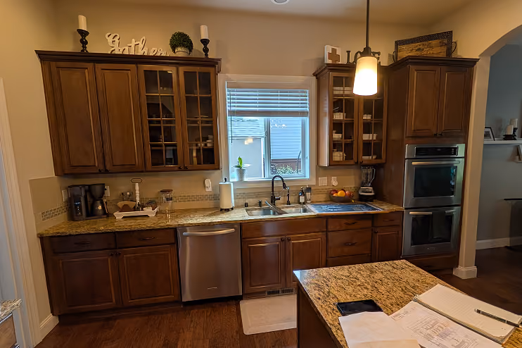 Modern kitchen with dark wood cabinets and a granite countertop, featuring a stainless steel dishwasher, double wall ovens, a double sink under a window, and an island with papers, a phone, and a pen on top.