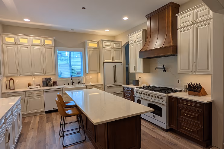 Modern kitchen with white and dark wood cabinets, a large island with beige countertop, two brown leather bar stools, stainless steel gas stove with a large wooden hood, a white refrigerator, and a window above the sink letting in natural light.