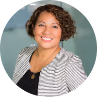 Smiling woman with short curly hair wearing a striped blazer and black top against a blurred background.