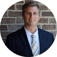 Smiling middle-aged man wearing a black suit and striped tie standing in front of a brick wall.