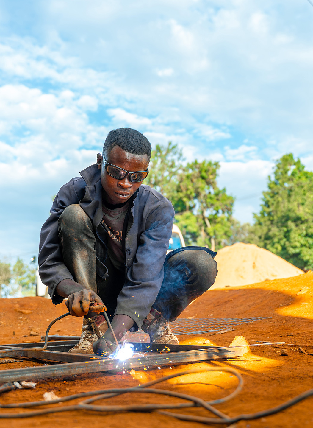 Alt: "Young man wearing safety glasses welding metal at a construction site in Kenya, sparks flying from the weld