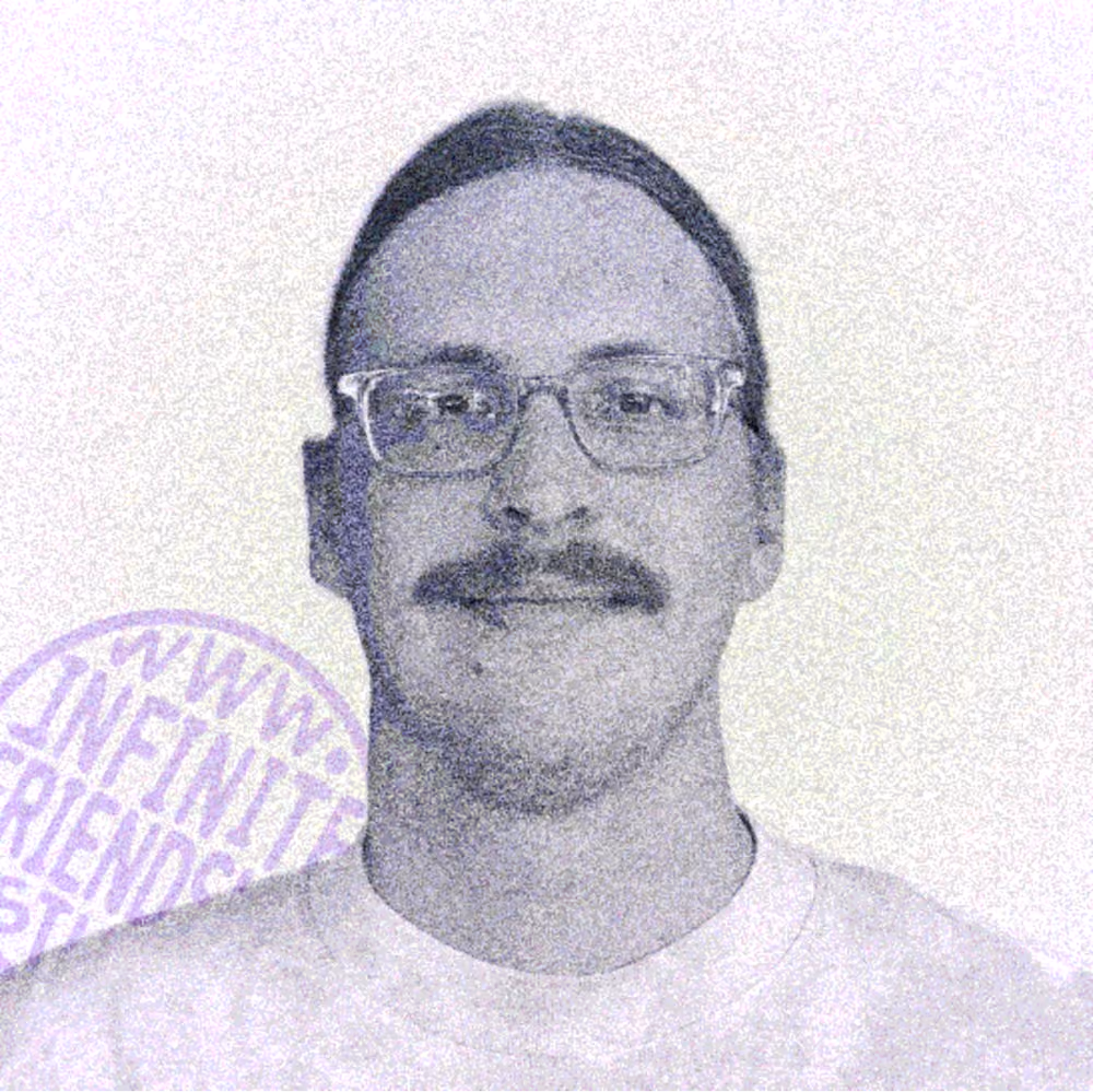 Headshot portrait of Will Halcomb, a man with pulled back long hair clear eyeglasses, a mustache and a white t shirt, standing in front of a white backdrop. There is a stamp for www.infinitefriendship.studio behind the man, advertising his boutique design and development studio.