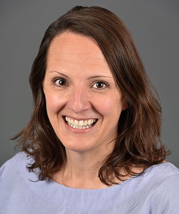 Smiling woman with shoulder-length brown hair wearing a light purple top on gray background.
