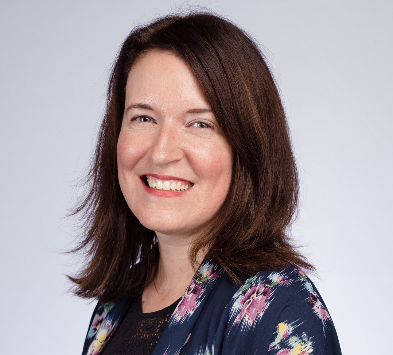 Woman with shoulder-length brown hair smiling, wearing a dark floral jacket and black top.