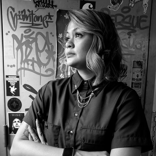 Black and white photo of a woman with crossed arms looking to the side against a graffiti wall.