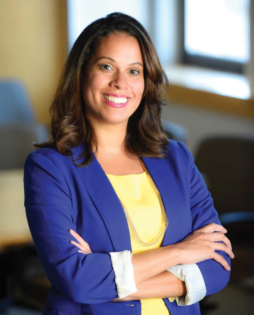 Smiling woman with brown hair wearing a blue blazer and yellow top, arms crossed.