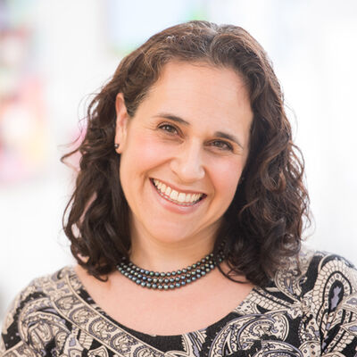 Smiling woman with curly dark hair wearing a patterned top and black beaded necklace.