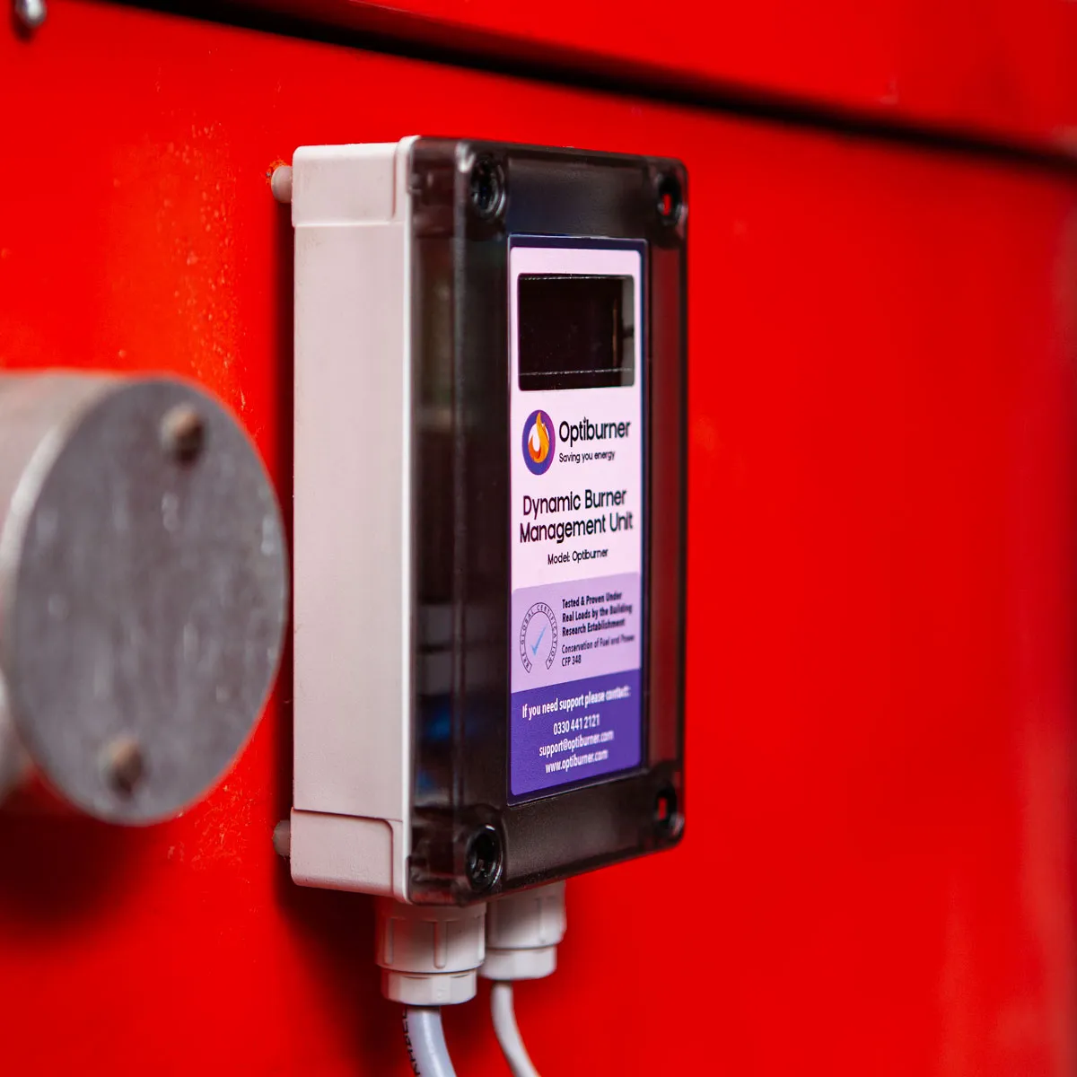 Optiburner Dynamic Burner Management Unit mounted on a red surface with two cables connected at the bottom.