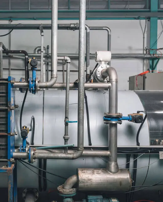 Close-up of an industrial boiler system with metal pipes, valves, and a cylindrical tank in a factory setting.