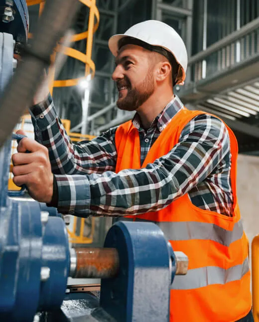 Male industrial worker in a white hard hat and orange safety vest operating machinery in a factory setting.