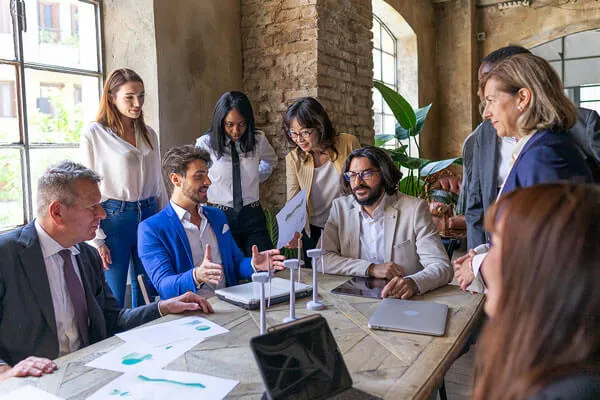 Seven diverse business professionals gathered around a table discussing a project with documents and laptops in a bright office.
