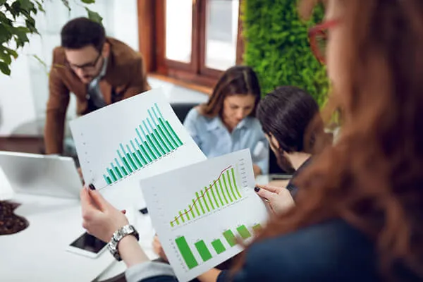 Person holding two sheets of paper with green and blue bar and line graphs in an office setting with three colleagues working in background.