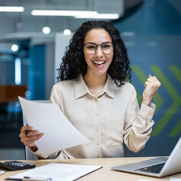 Smiling woman with curly hair and glasses celebrating while holding a document in an office setting.