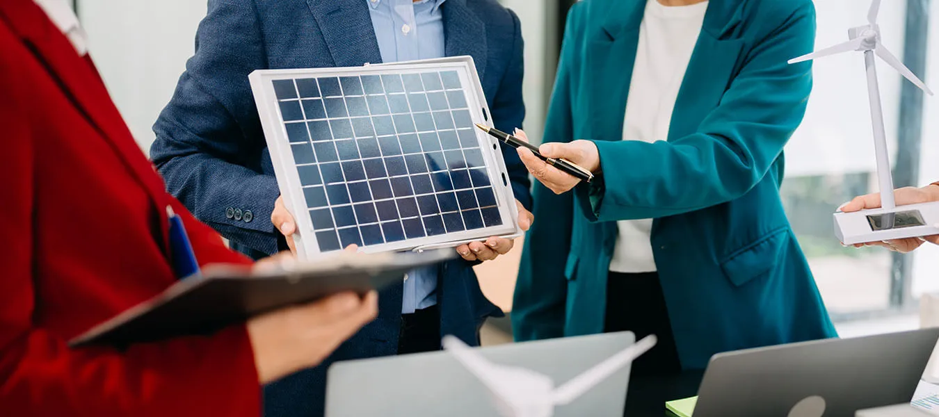 Three professionals discussing renewable energy solutions, one holding a solar panel, another pointing at it with a pen, and a third holding a small wind turbine model.