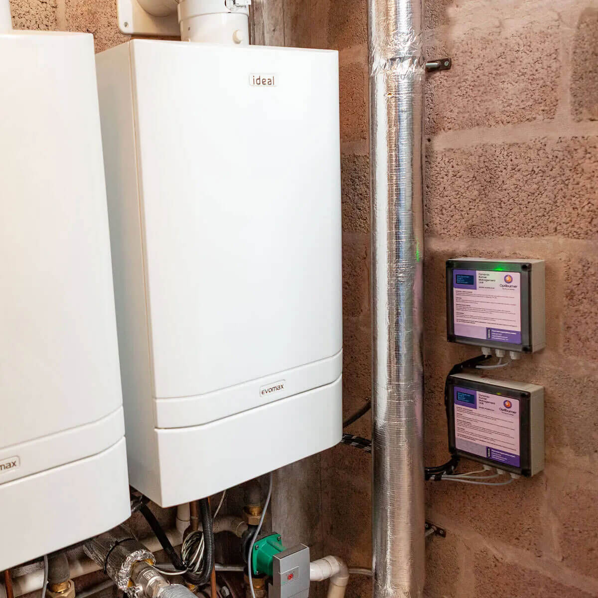 White Ideal Evomax boiler unit installed on a brick wall next to metal insulated pipes and two mounted electronic control boxes.