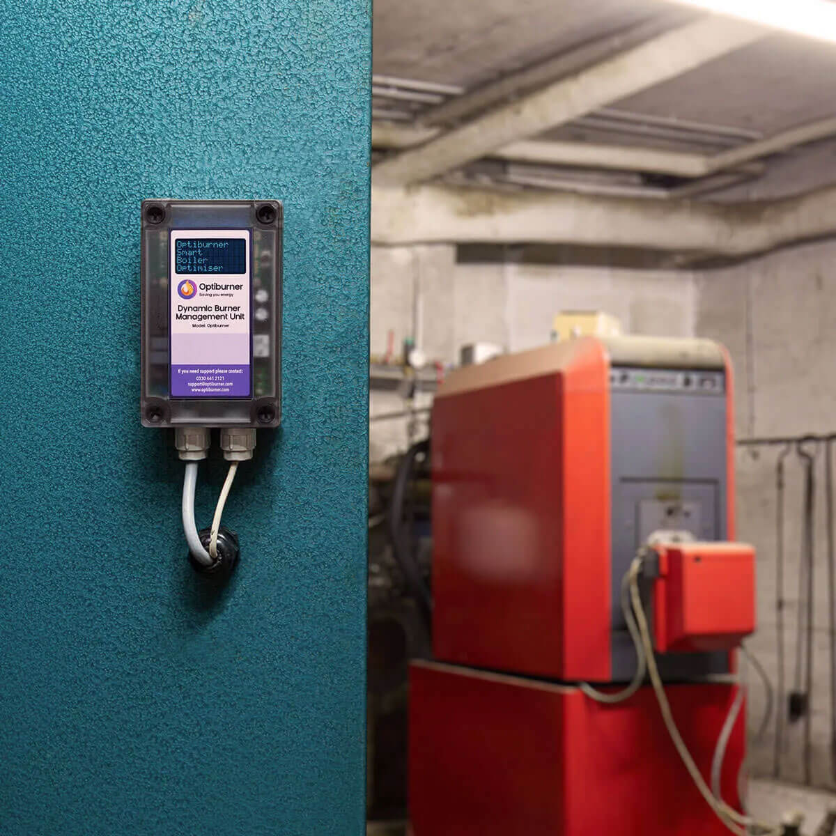 Optiburner's Dynamic Burner Management Unit mounted on a textured teal wall with a blurred industrial boiler in the background.
