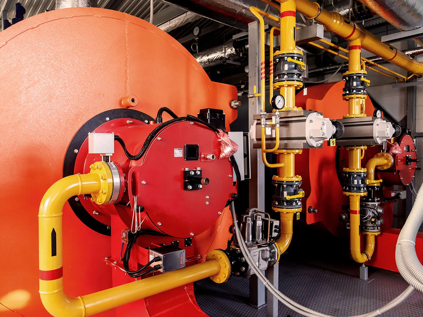 Industrial boilers with bright orange bodies and connected yellow pipes inside a mechanical room.