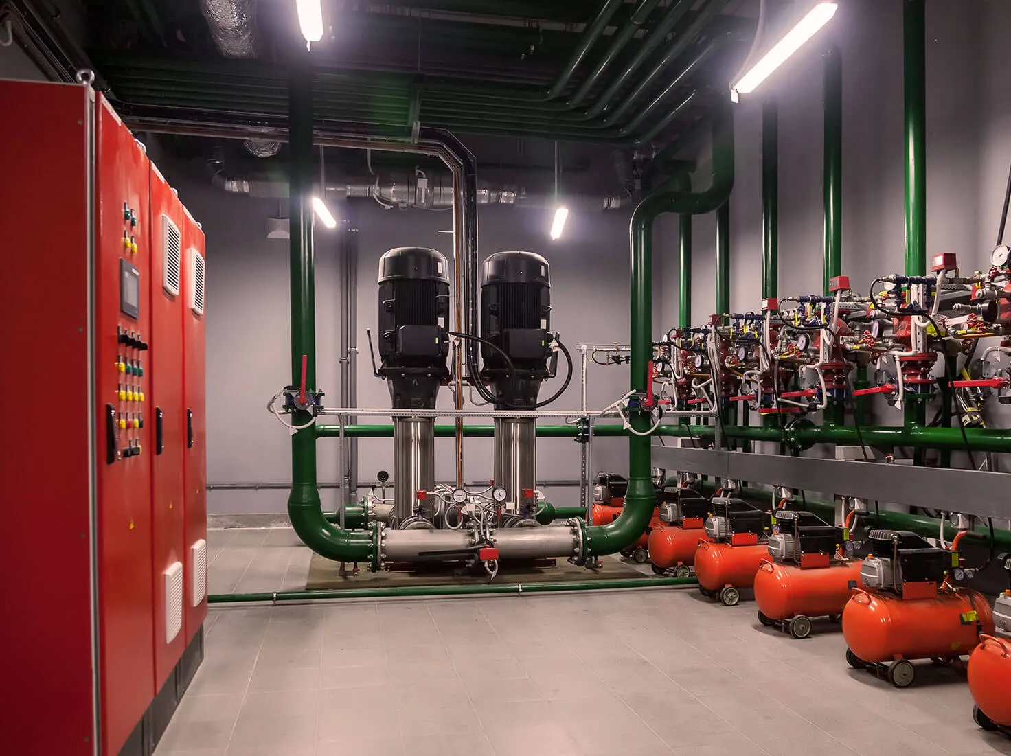 Industrial boiler room with green pipes, two large black pumps, red compressor tanks, and red control panels on the left.