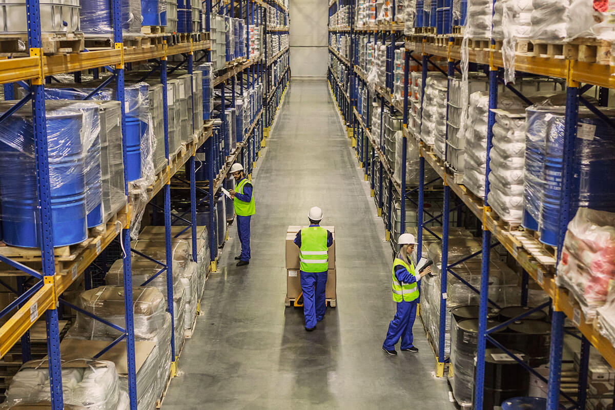 Warehouse aisle with high shelves stocked with pallets and barrels, and three workers wearing safety vests and helmets managing inventory.