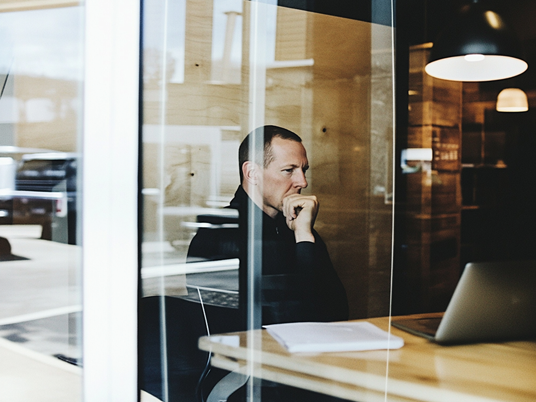 Man thinking in front of a laptop, seen through a café window