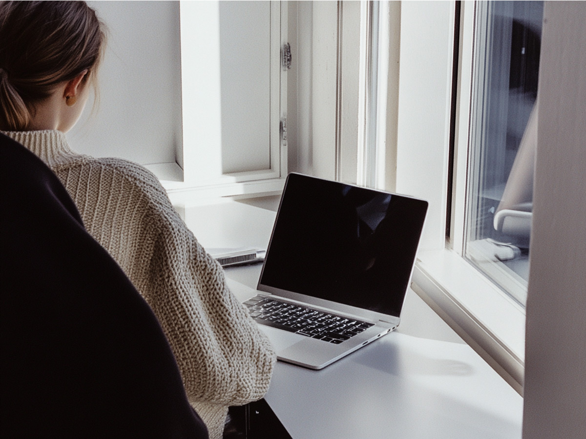 Woman in a knit sweater working on a laptop by a window
