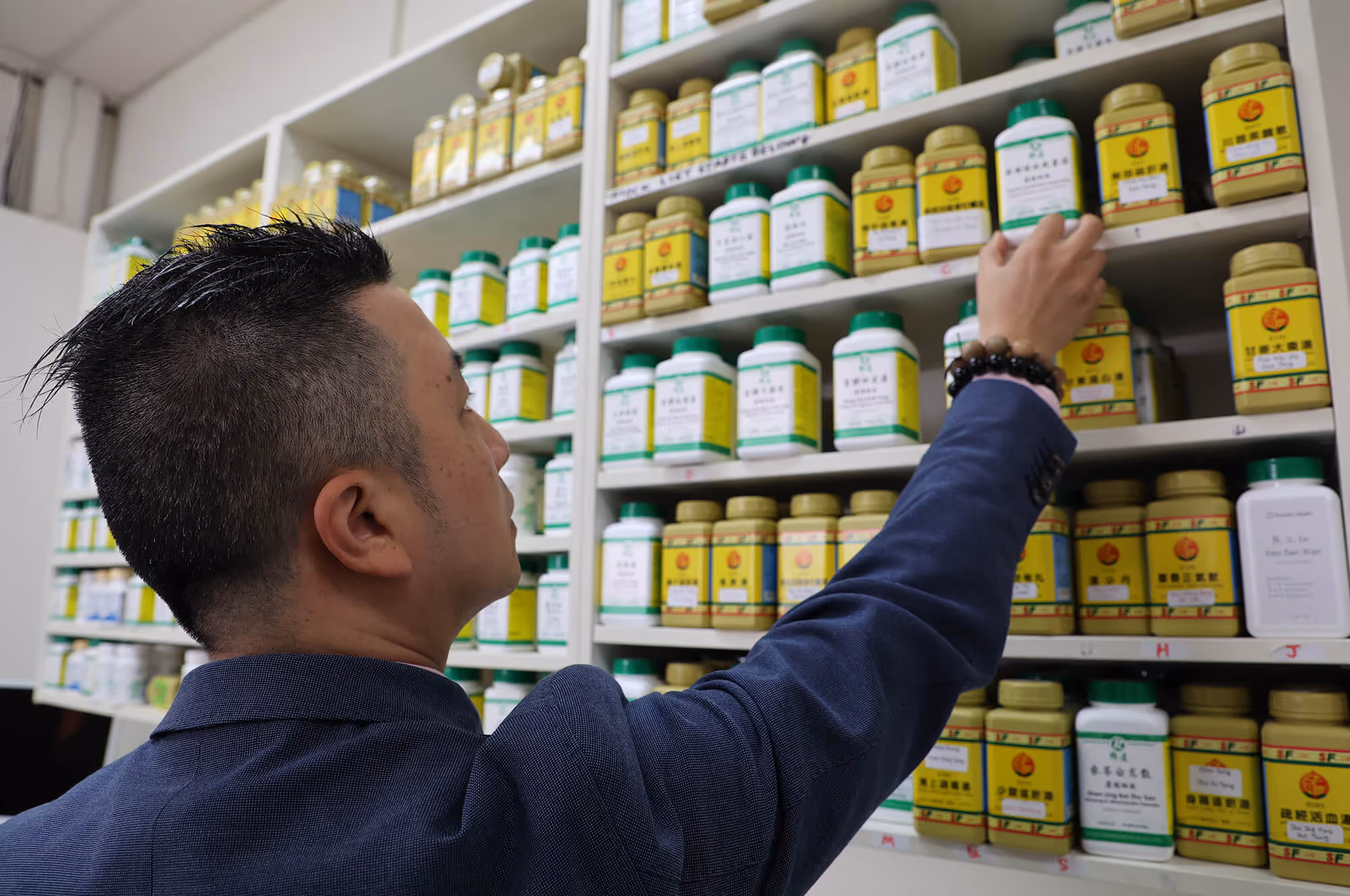 Man in a navy blazer reaching for a container on a shelf filled with green and yellow traditional medicine bottles.