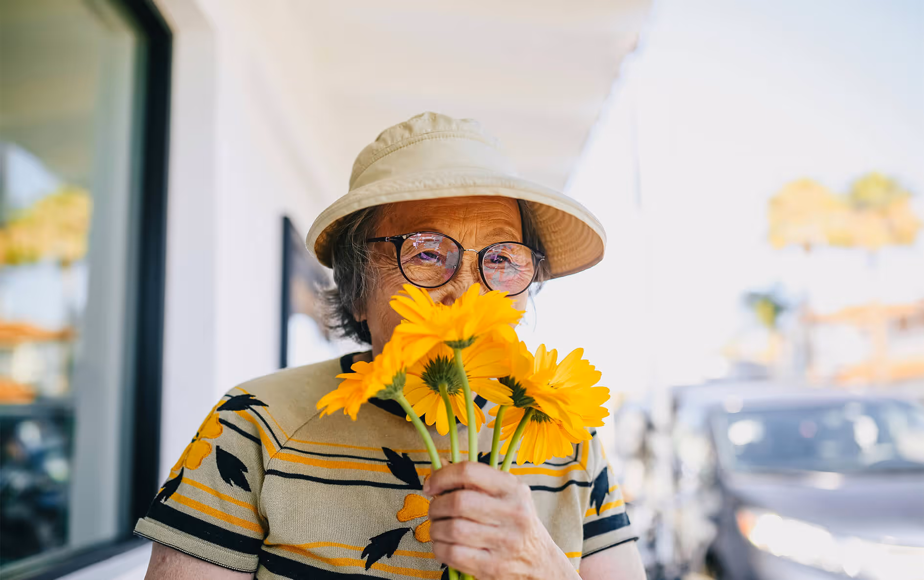 Elderly person wearing glasses and a sun hat smelling a bouquet of yellow flowers outside.