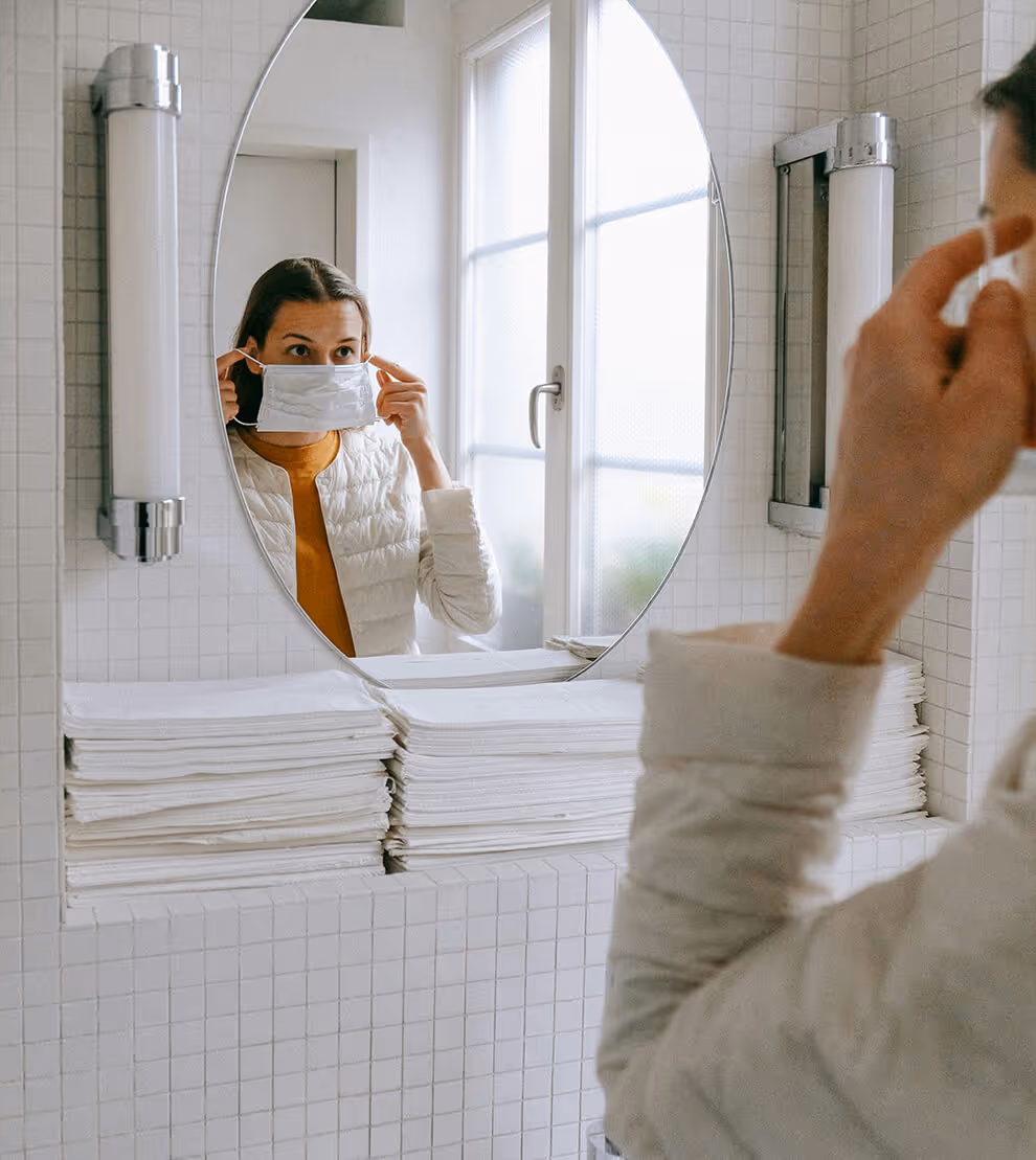 Person wearing a white jacket putting on a white face mask while looking into a round bathroom mirror.
