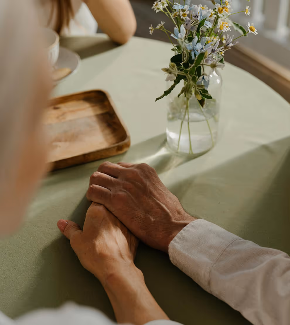 Two people holding hands on a green tablecloth next to a glass vase with small white and blue flowers.