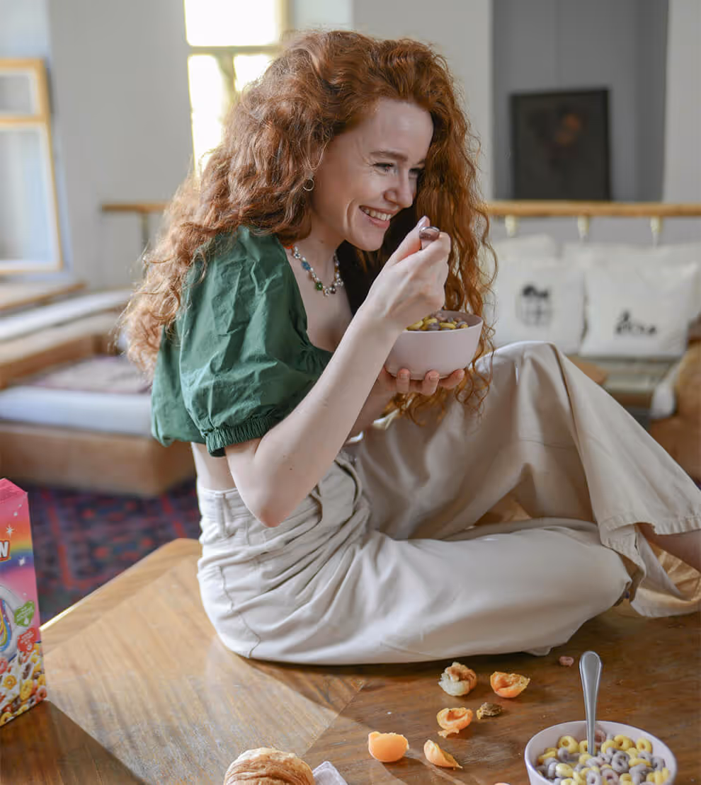 Smiling woman with curly red hair sitting on a table eating colorful cereal from a bowl with a spoon.