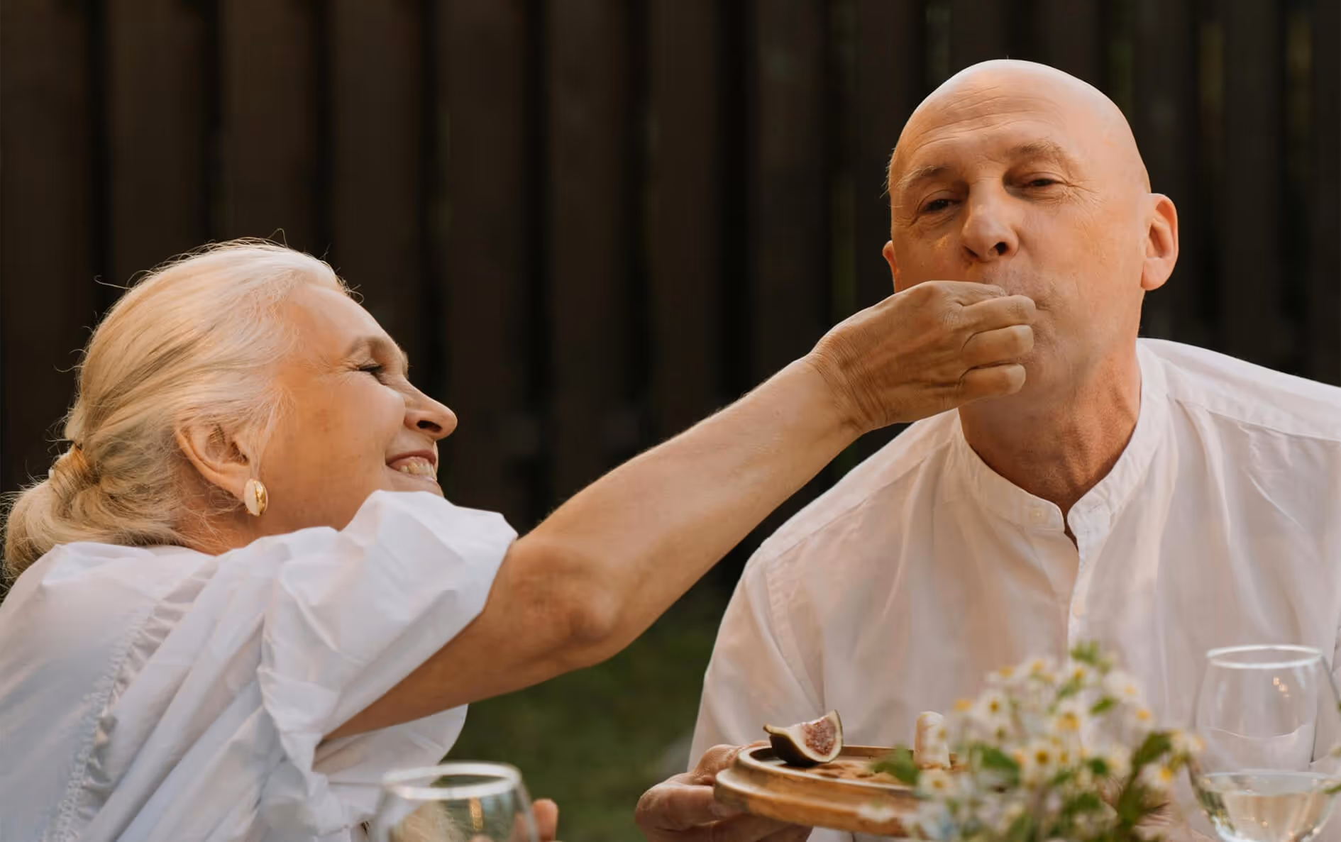 Elderly woman feeding a bite of food to an elderly man at a table outdoors.