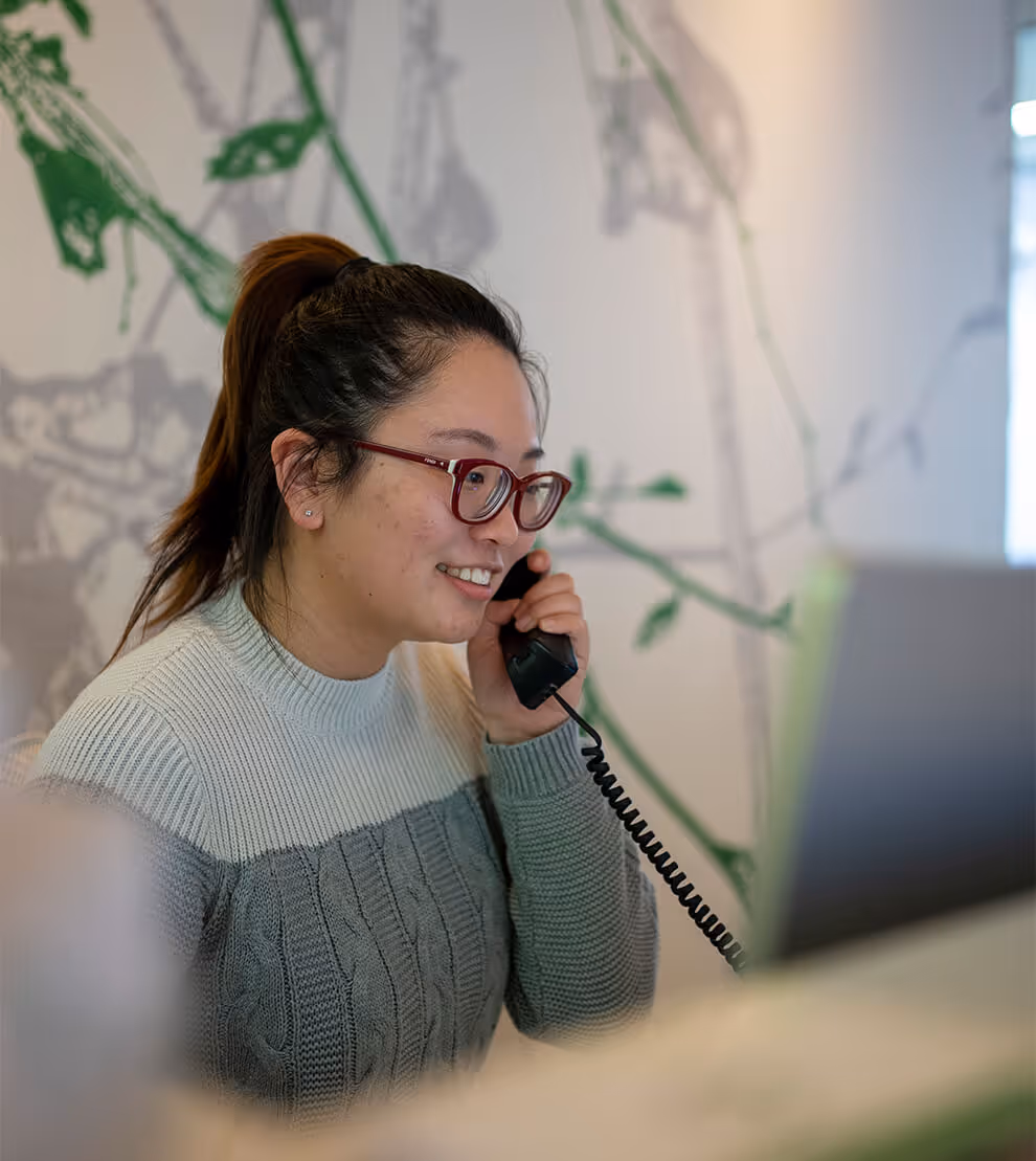 Woman with glasses and a ponytail smiling and speaking on a corded phone at a desk with a laptop.