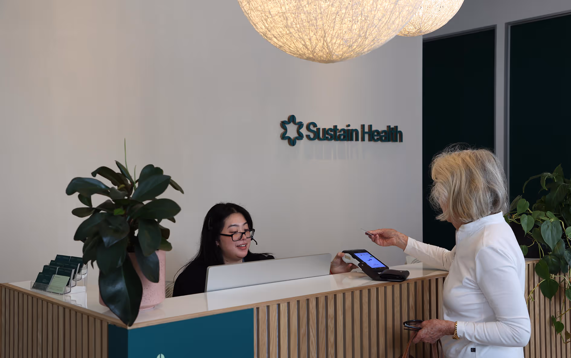 Receptionist at Sustain Health desk with headset helping an elderly woman making a payment with a card on a contactless machine.