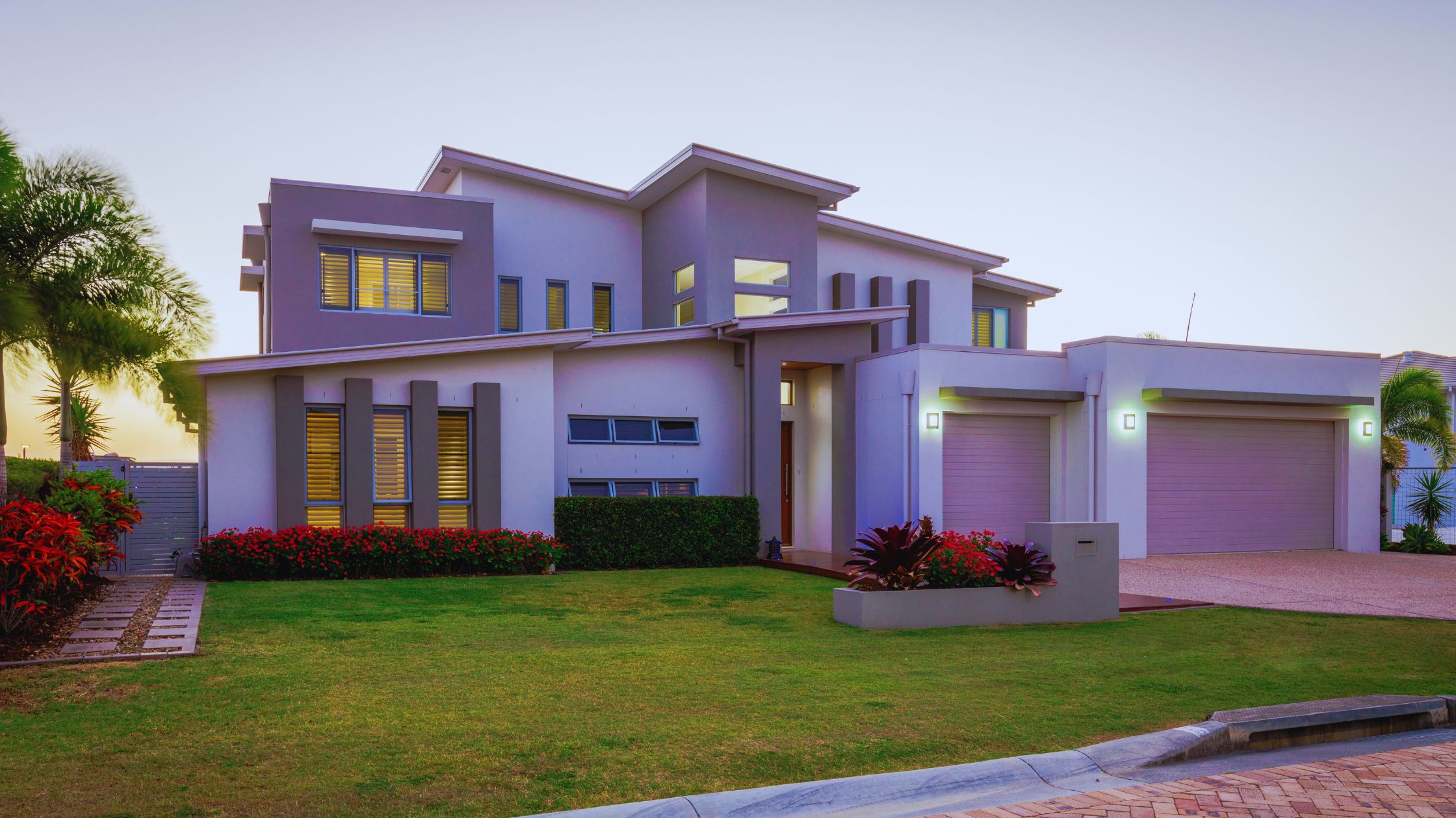 "A modern suburban home with a spacious driveway and double garage, set against a vibrant sunset sky, featuring well-maintained landscaping and a cozy porch light illuminating the entryway.