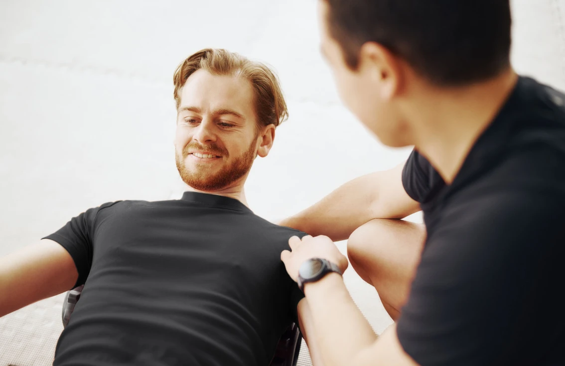 Personal trainer assisting a man with a core exercise on a mat in a bright, minimal indoor setting.